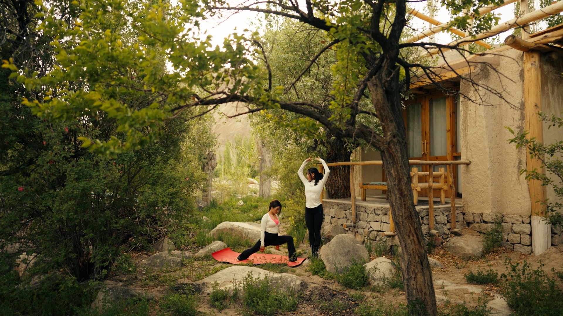Two people doing yoga together in a lush garden beside a traditional Ladakhi building